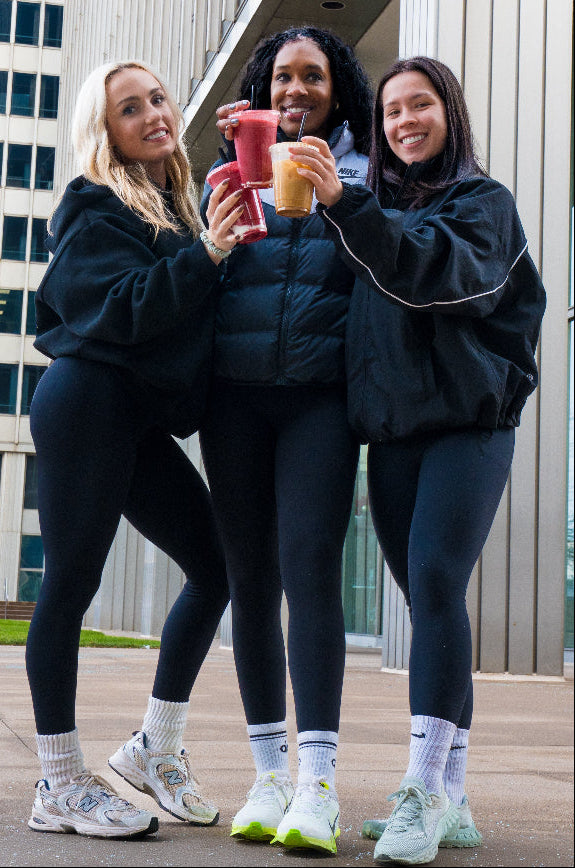 Three women standing outdoors in front of a modern building, wearing matching black athletic outfits and holding colorful smoothie cups.