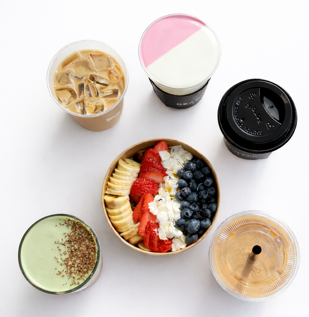 An overhead flat-lay of six beverage and food items on a clean white background: an iced coffee, a pink-and-white drink, a black-topped takeaway coffee cup, a green matcha drink with sprinkles, a fruit and cream acai bowl, and a tan-colored drink in a clear cup.